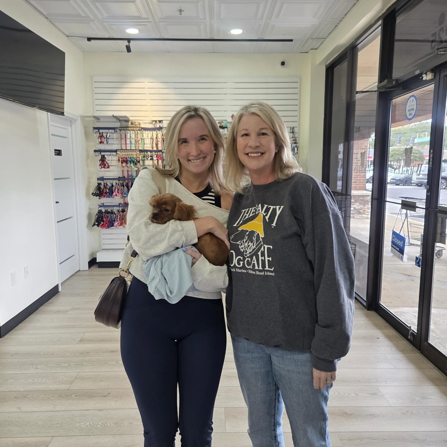 Two women smiling together while one holds a brown puppy inside the boutique